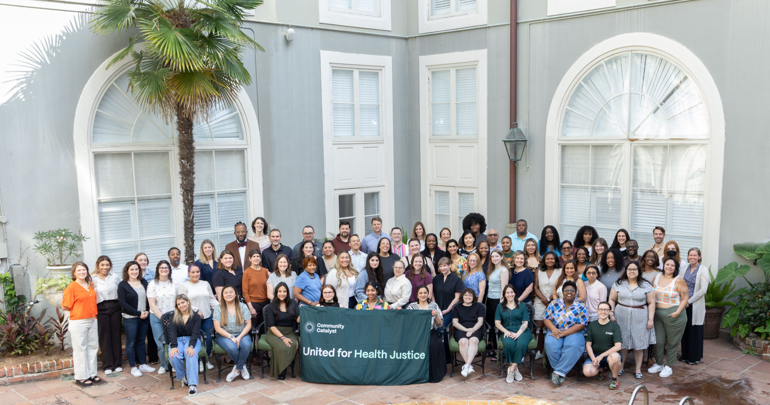 Community Catalyst staff pose for a photo in front of large windows and a palm tree. In the middle of the group is a dark green banner with Community Catalyst's logo and the words "United for Health Justice" written.