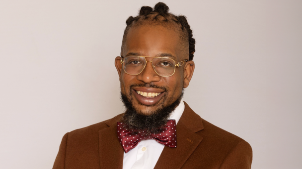This portrait features a man smiling with braided locs, gold rimmed glasses. He is wearing a brown suit, white dress shirt, and a red and white bowtie.