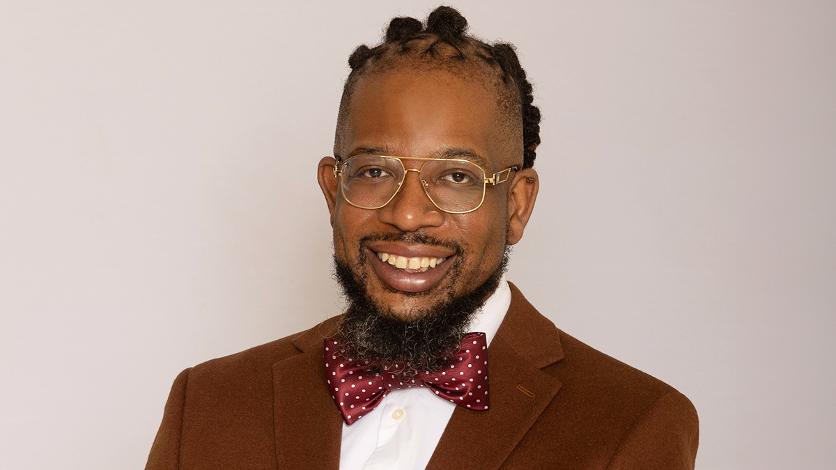This portrait features a man smiling with braided locs, gold rimmed glasses. He is wearing a brown suit, white dress shirt, and a red and white bowtie.