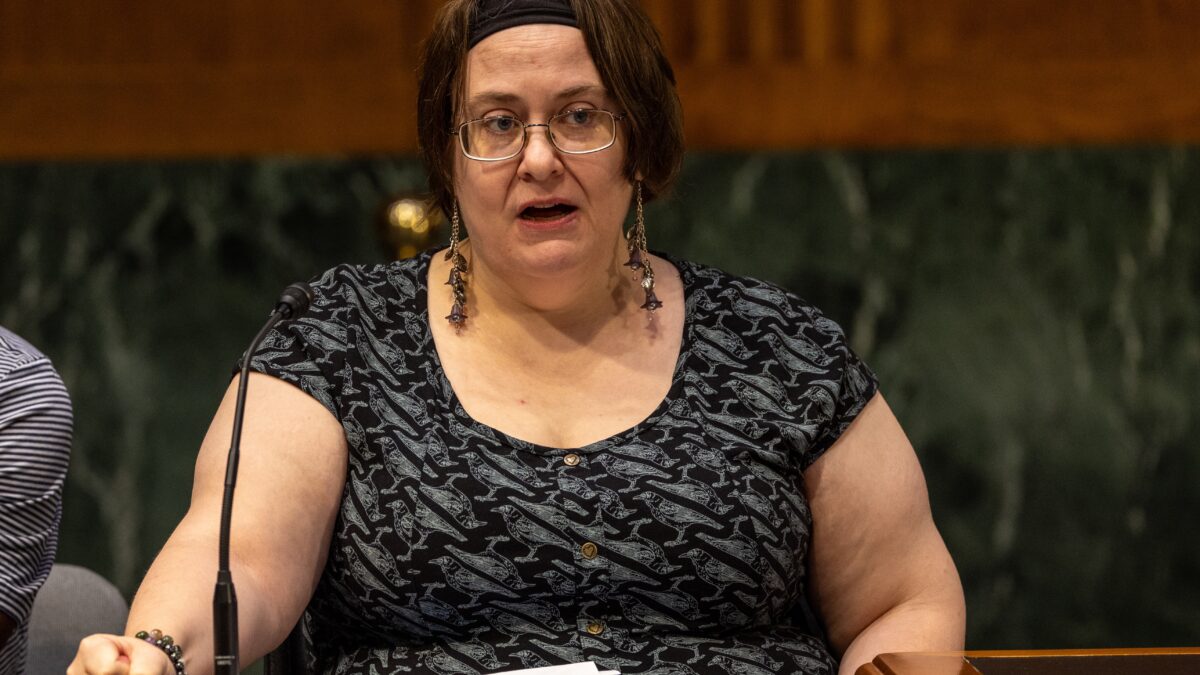 Person speaks into a microphone at a roundtable in Washington, D.C.. She is wearing gold earrings, a black headband, and a black and grey shirt with a bird graphic. There are are printed documents and a cellphone on a wooden table in front of the speaker