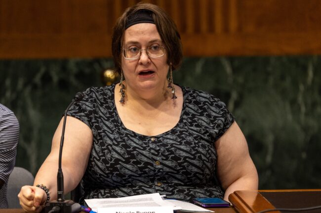 Person speaks into a microphone at a roundtable in Washington, D.C.. She is wearing gold earrings, a black headband, and a black and grey shirt with a bird graphic. There are are printed documents and a cellphone on a wooden table in front of the speaker