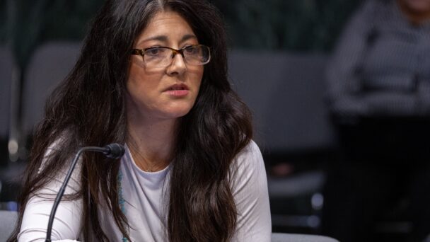 A storyteller with long dark hair and wearing a white long-sleeved shirt with glasses speaks at a roundtable.
