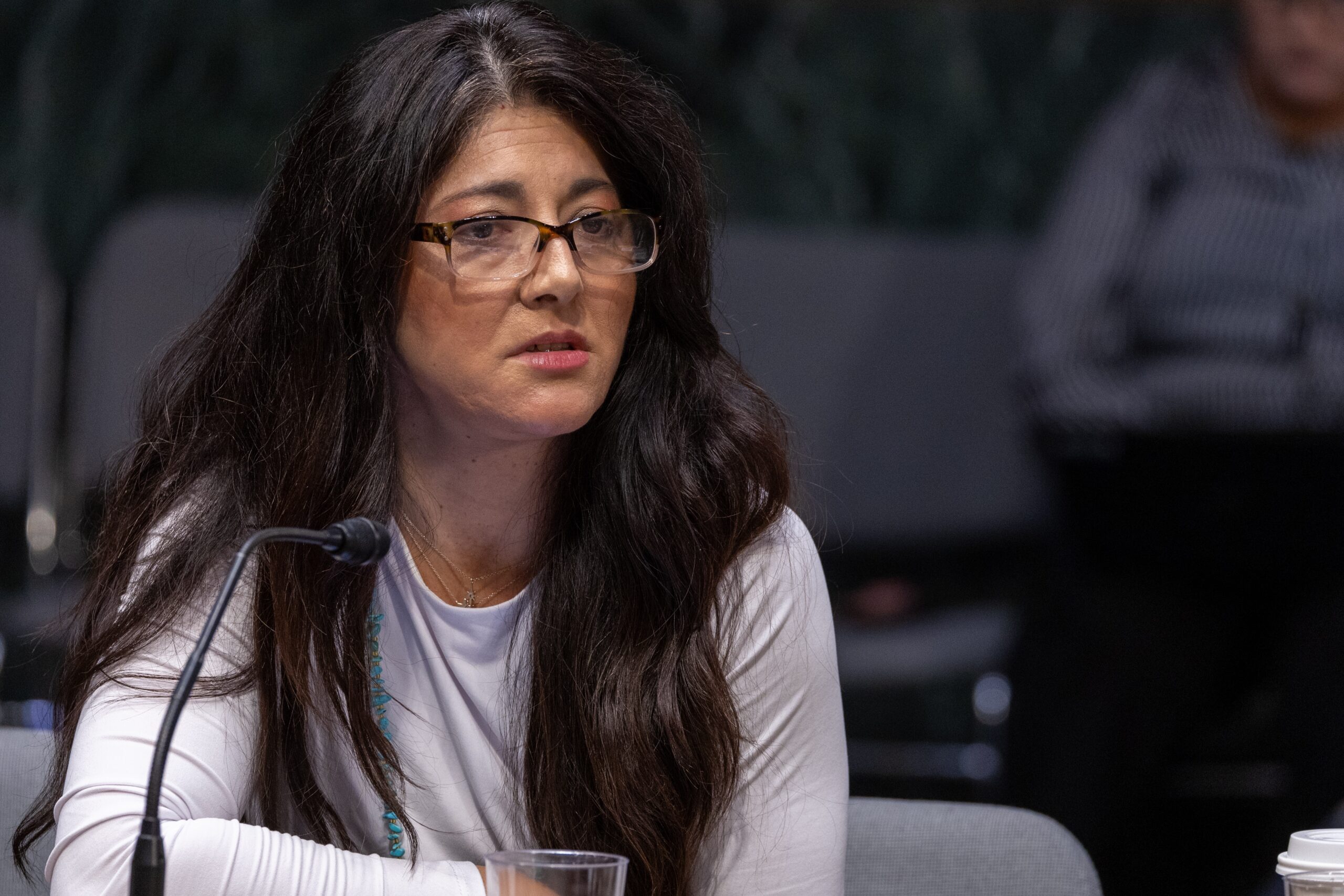 A storyteller with long dark hair and wearing a white long-sleeved shirt with glasses speaks at a roundtable.