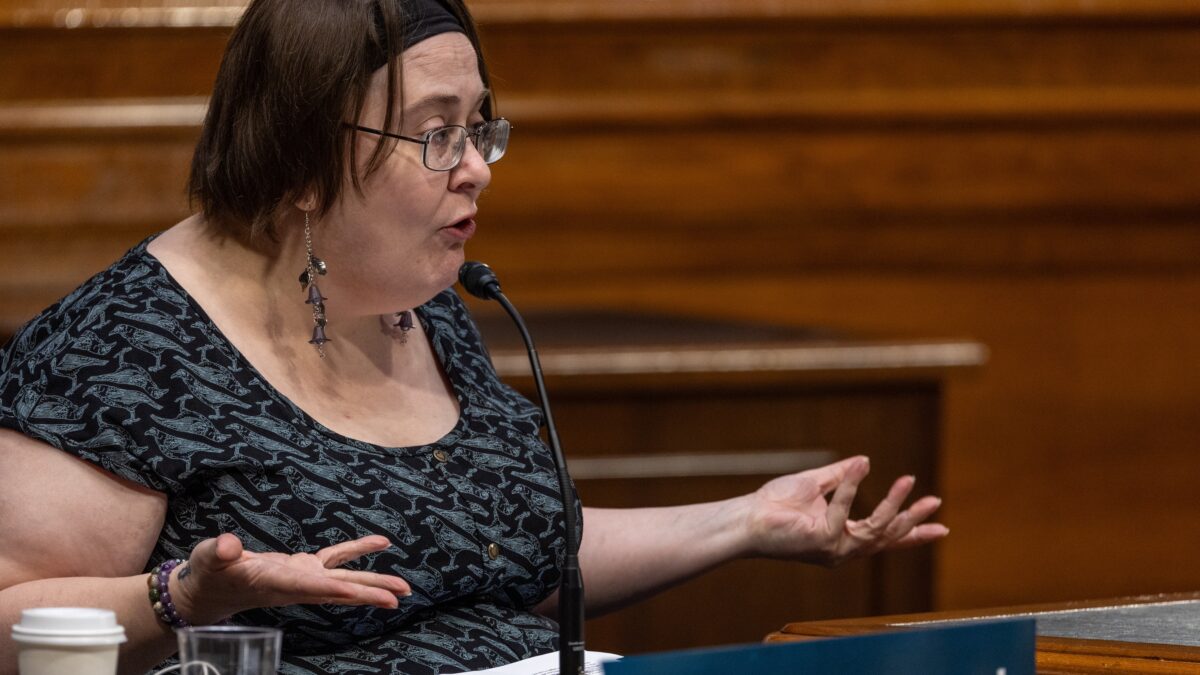 Person speaks into a microphone at a roundtable in Washington, D.C.. She is wearing earrings, a black headband, and a black and grey shirt with bird graphics. There are are printed documents and a cellphone on a wooden table in front of the speaker.