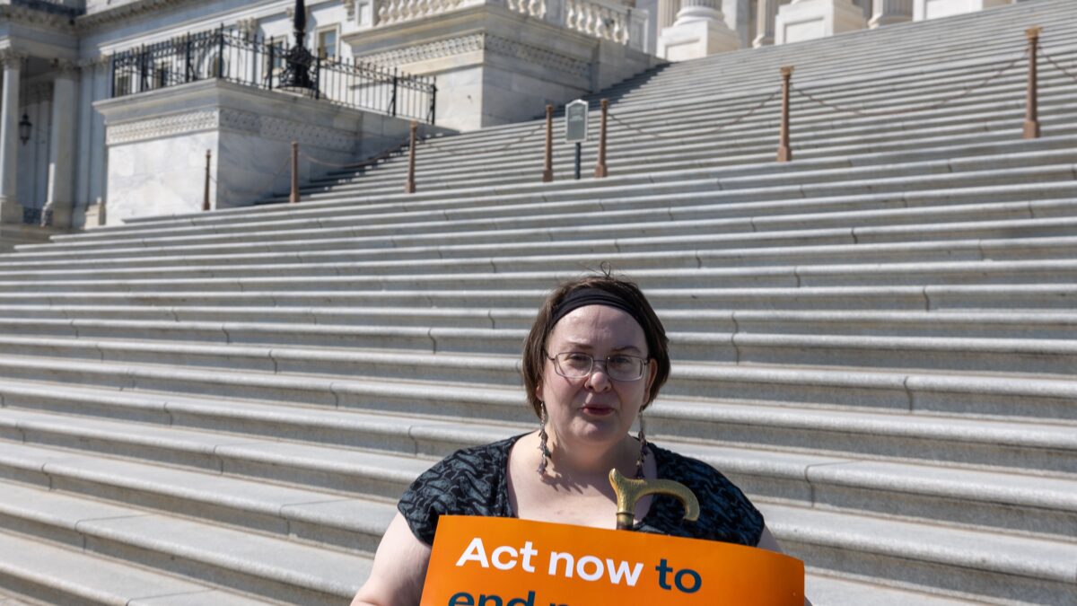 A storyteller sits in a wheelchair in front of the steps of the U.S. capitol and holds a sign that says "Act now to end medical debt."