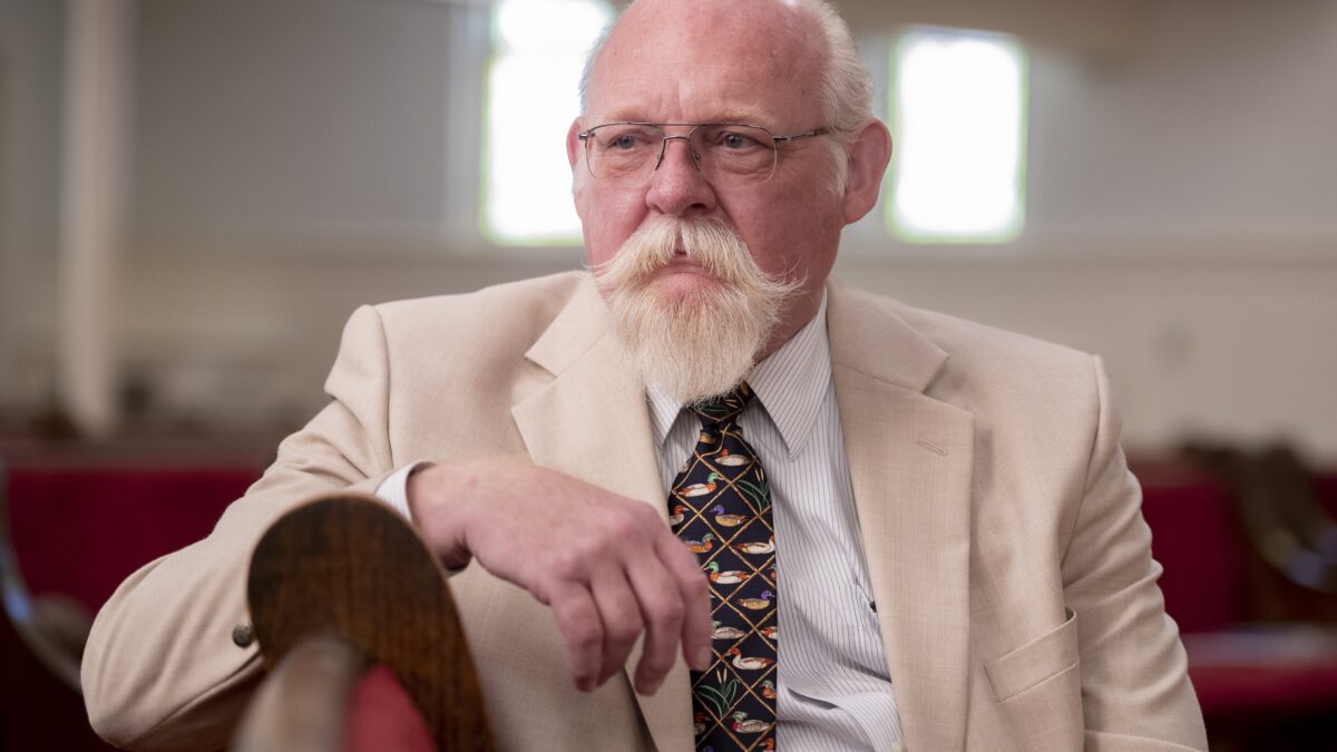 A pastor sits on a pew in a church. The pastor is wearing a tan suit with a dark multicolored tie.