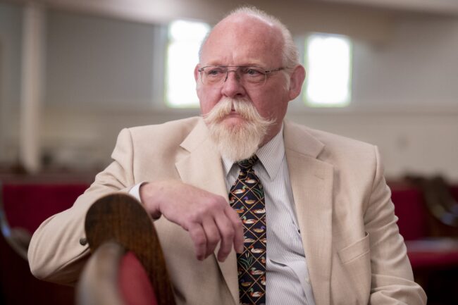 A pastor sits on a pew in a church. The pastor is wearing a tan suit with a dark multicolored tie.