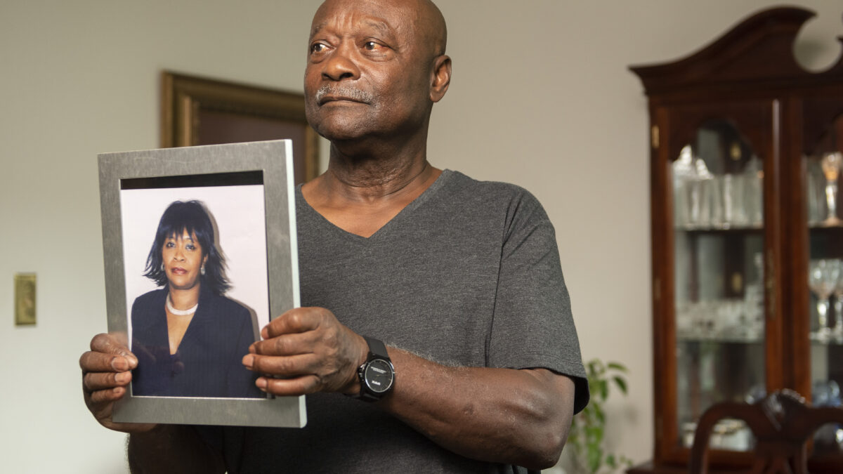 A storyteller holds a framed photo of his late wife while standing in his living room. He is wearing a grey shirt.