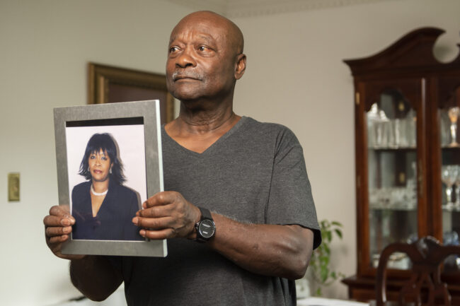 A storyteller holds a framed photo of his late wife while standing in his living room. He is wearing a grey shirt.