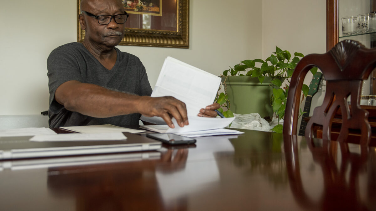 A storyteller sits at a wooden dining table and combs through several pages of medical bills and other hospital documents. He is wearing a grey shirt and glasses.