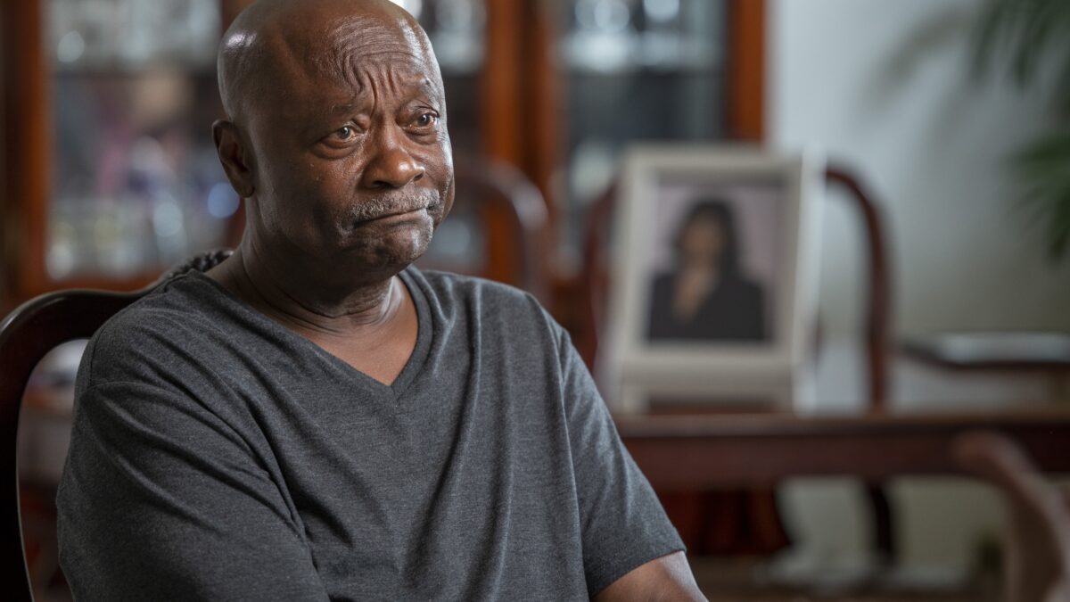 A storyteller sits in a living room with a dining table and framed photo of his late wife in the background. He is wearing a grey shirt.