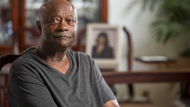 A storyteller sits in a living room with a dining table and framed photo of his late wife in the background. He is wearing a grey shirt.