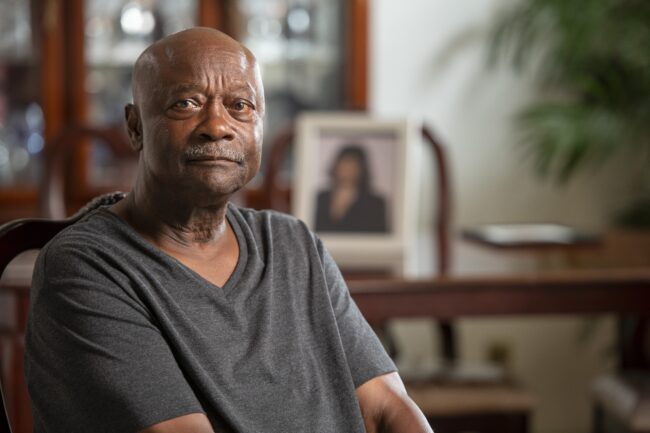 A storyteller sits in a living room with a dining table and framed photo of his late wife in the background. He is wearing a grey shirt.