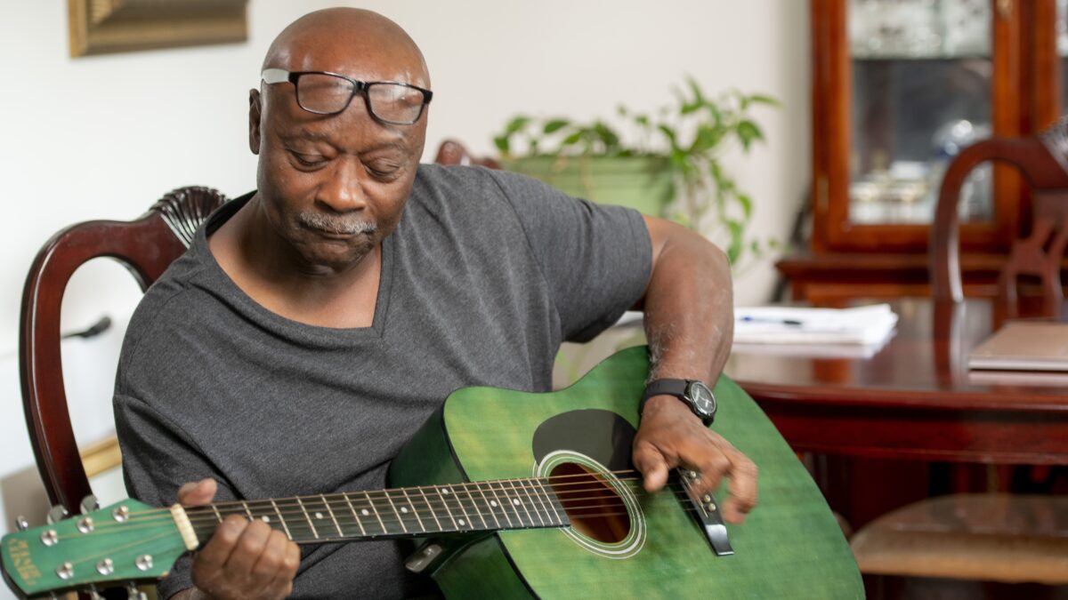 A storyteller wearing glasses and a grey shirt plays a green guitar. There are medical bills stacked on a wooden dining table in the background.