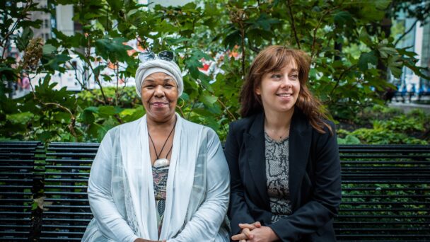 An older person and younger person sit on a park bench in front of a leafy tree. The person on the left is dressed in a sheer white tunic, matching head wrap, and a beaded multicolored top with a stone necklace and sunglasses. The person on the right has brown hair and is wearing a necklace, black blazer and grey, and black top.