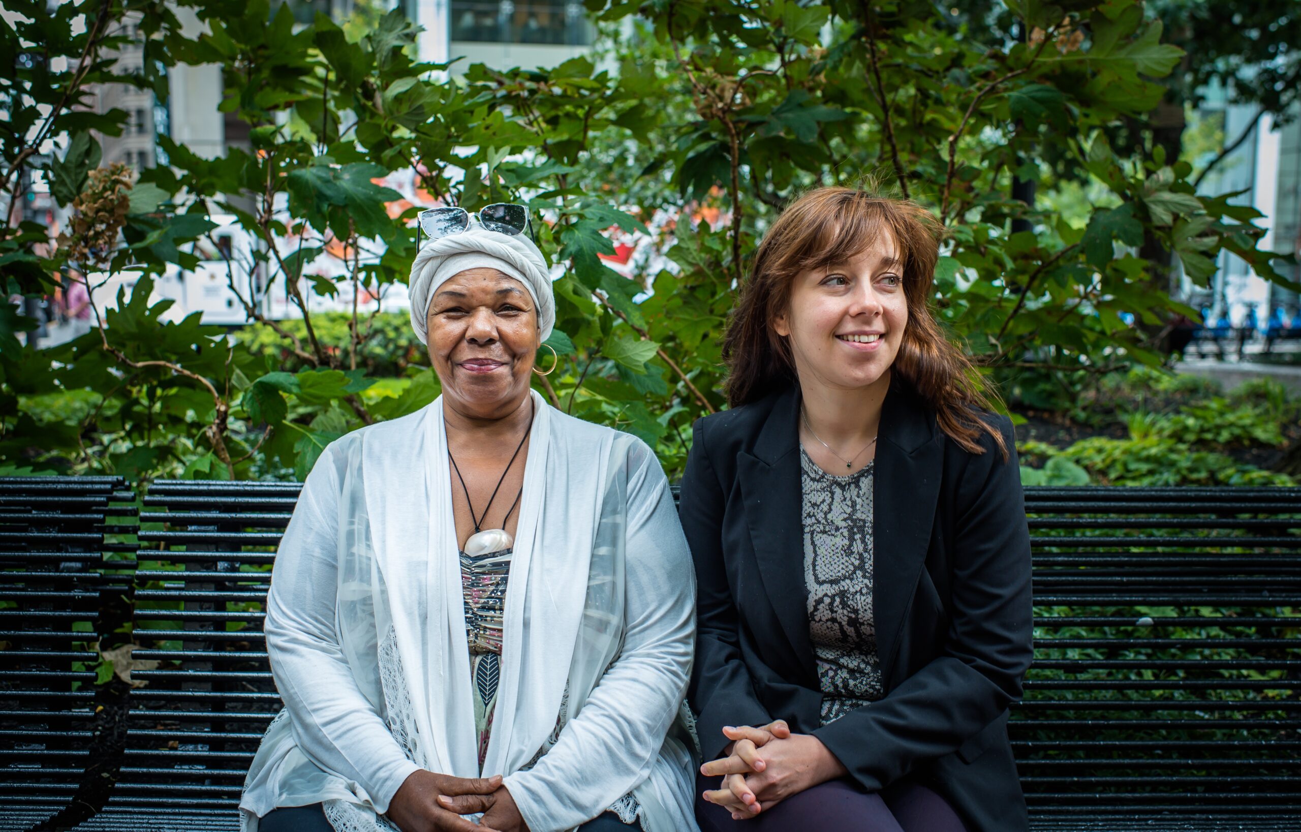 An older person and younger person sit on a park bench in front of a leafy tree. The person on the left is dressed in a sheer white tunic, matching head wrap, and a beaded multicolored top with a stone necklace and sunglasses. The person on the right has brown hair and is wearing a necklace, black blazer and grey, and black top.