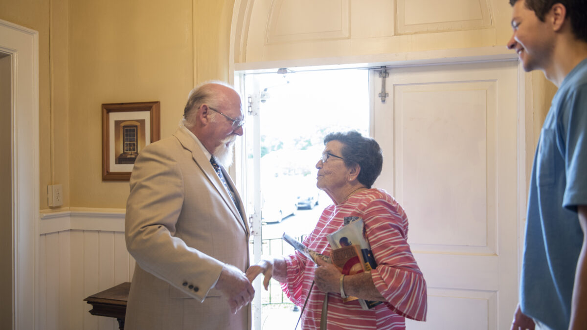 A pastor wearing glasses and a tan suit speaks to an elderly woman wearing glasses and a pink striped blouse in a church. She is holding documents and a purse. A third person smiles in the foreground in the photo.