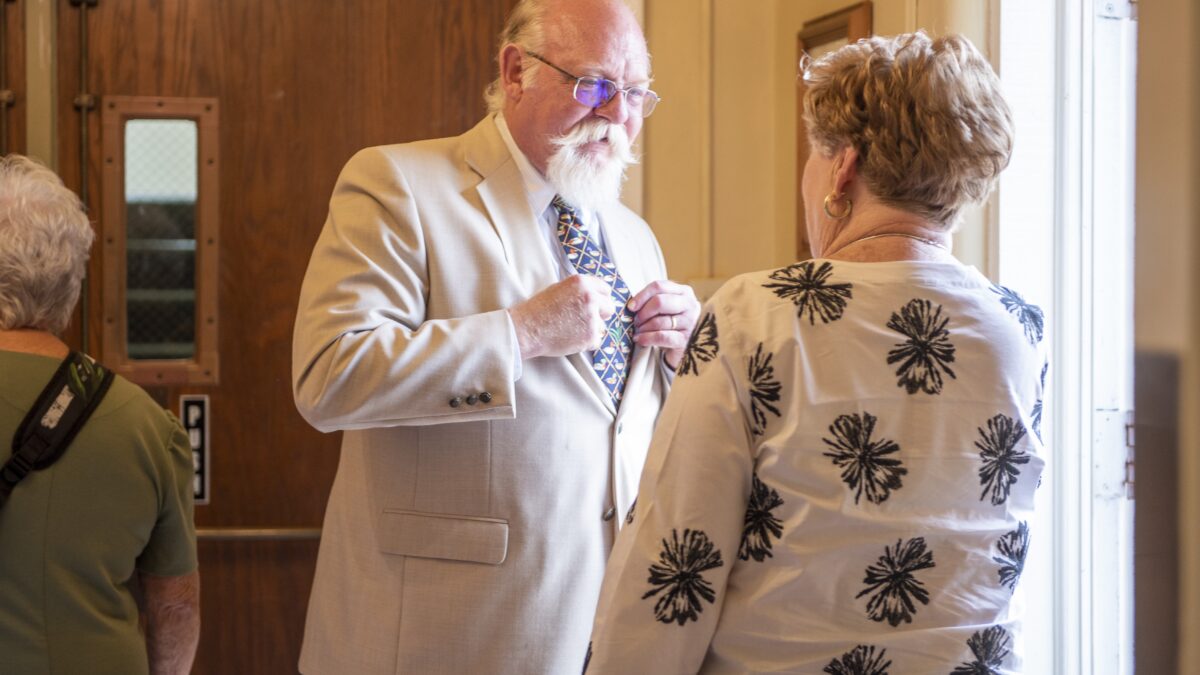 A paster wearing glasses, a multicolored tie, and a tan suit speaks to a church patron in front of a church door exit.