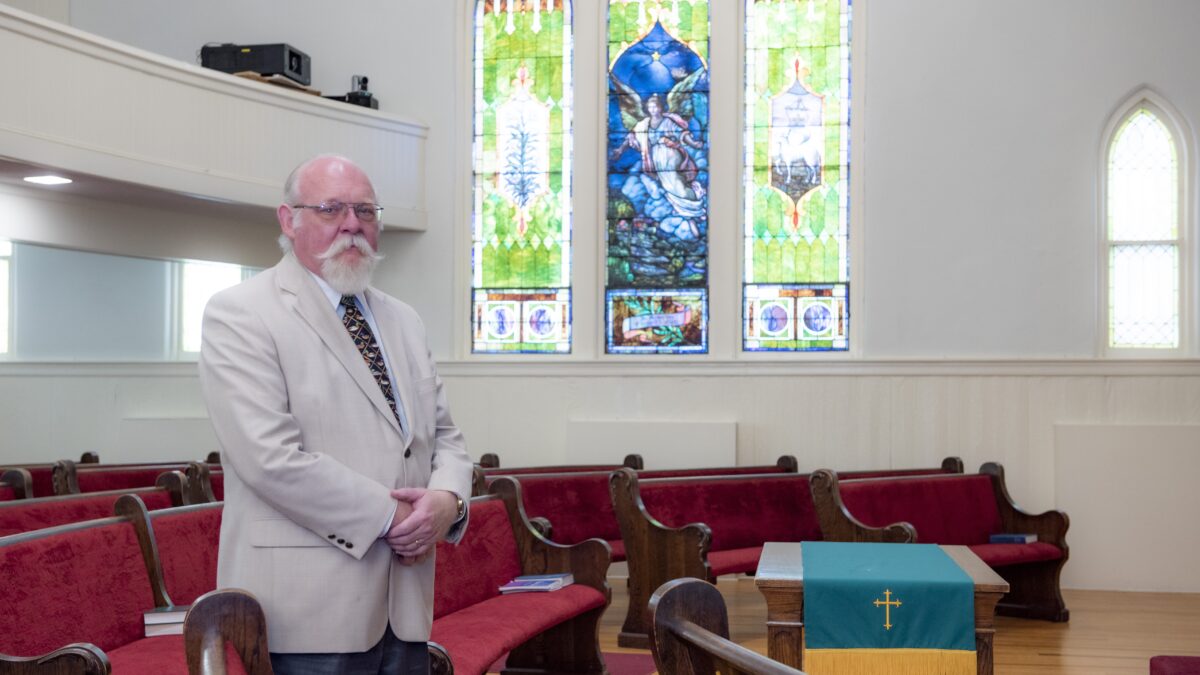 A pastor wearing glasses and a tan suit stands in front of church pews and stained glass windows