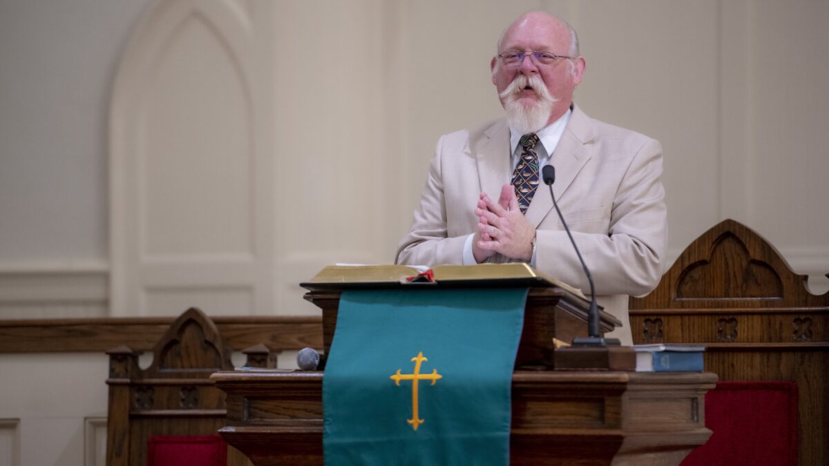 A pastor wearing glasses and a tan suit speaks at a pulpit in a church