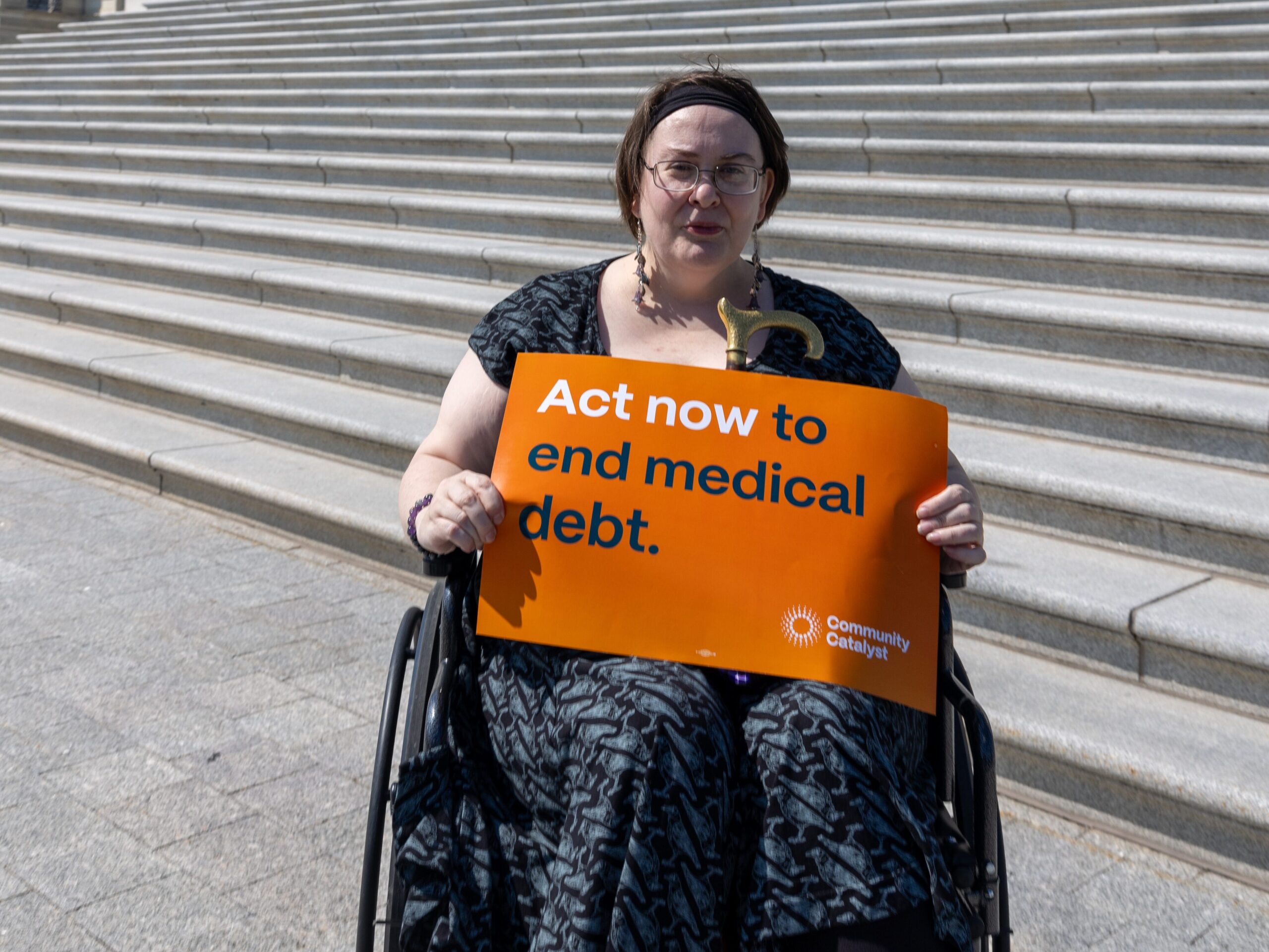 A storyteller sits in a wheelchair in front of the steps of the U.S. capitol and holds a sign that says "Act now to end medical debt."