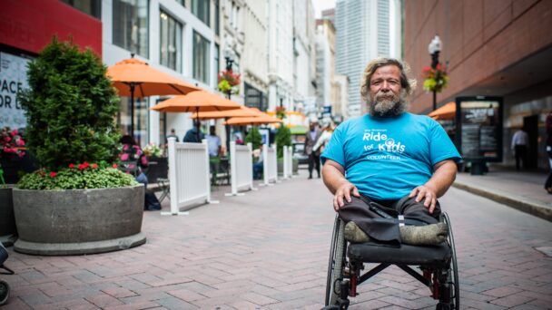 A person in a wheelchair wearing a blue t-shirt, black pants, and grey socks poses in a city center.