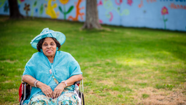 A woman smiles in a park while seated in her wheelchair. She is wearing a blue hat, a blue top and cardigan, and a blue skirt.