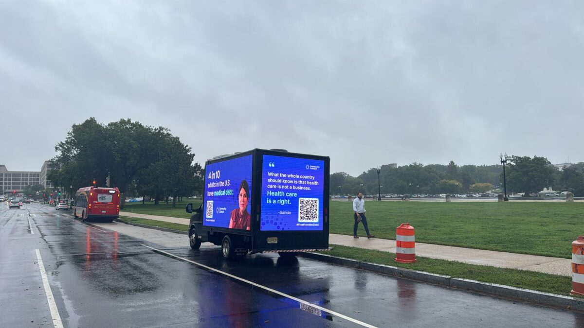 A mobile billboard is parked on a tree-lined road that reads "4 in 10 adults in the U.S. have medical debt" and "What the whole country should know is that health care is not a business. Health care is a right."