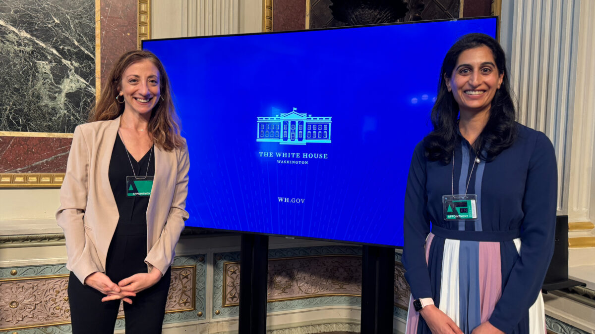 Two people pose in the White House. One person is wearing a beige blazer with black pants and the other person is wearing a multicolor skirt with a navy blouse. Behind them are ornate marble paintings.