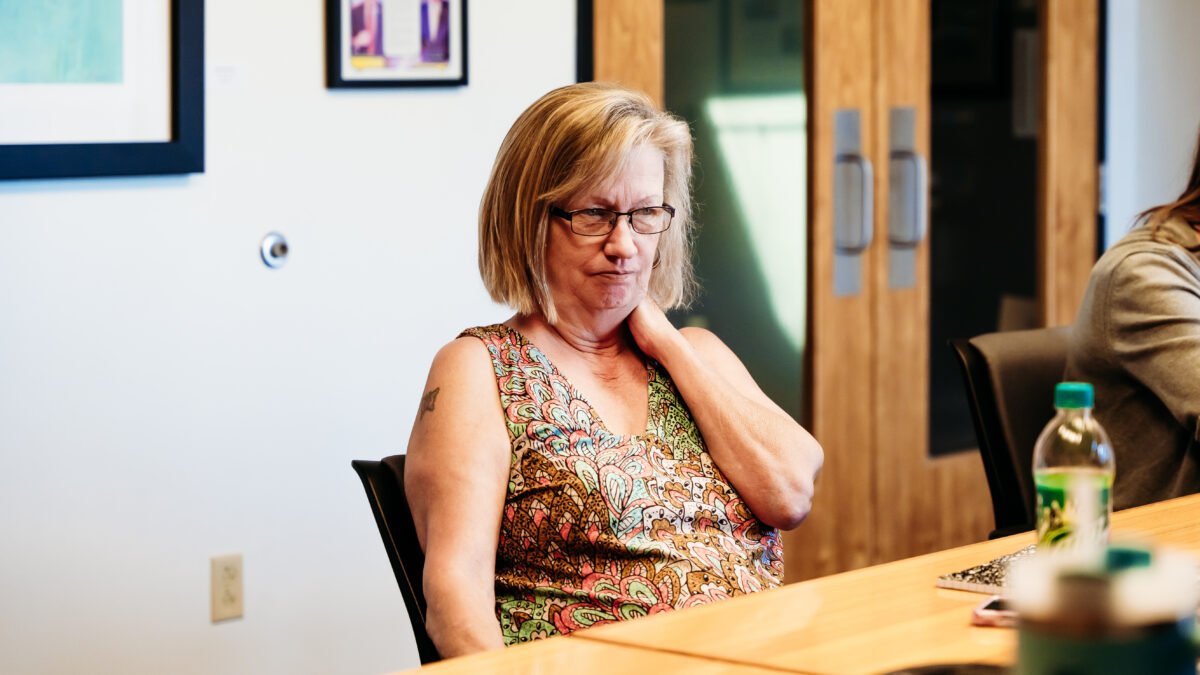 A person with short blonde hair and glasses looks ahead while sitting at a conference table.