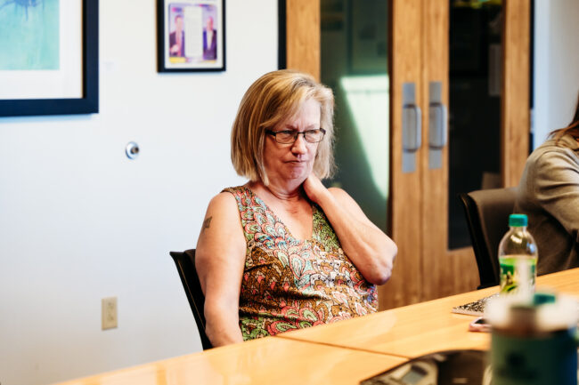 A person with short blonde hair and glasses looks ahead while sitting at a conference table.