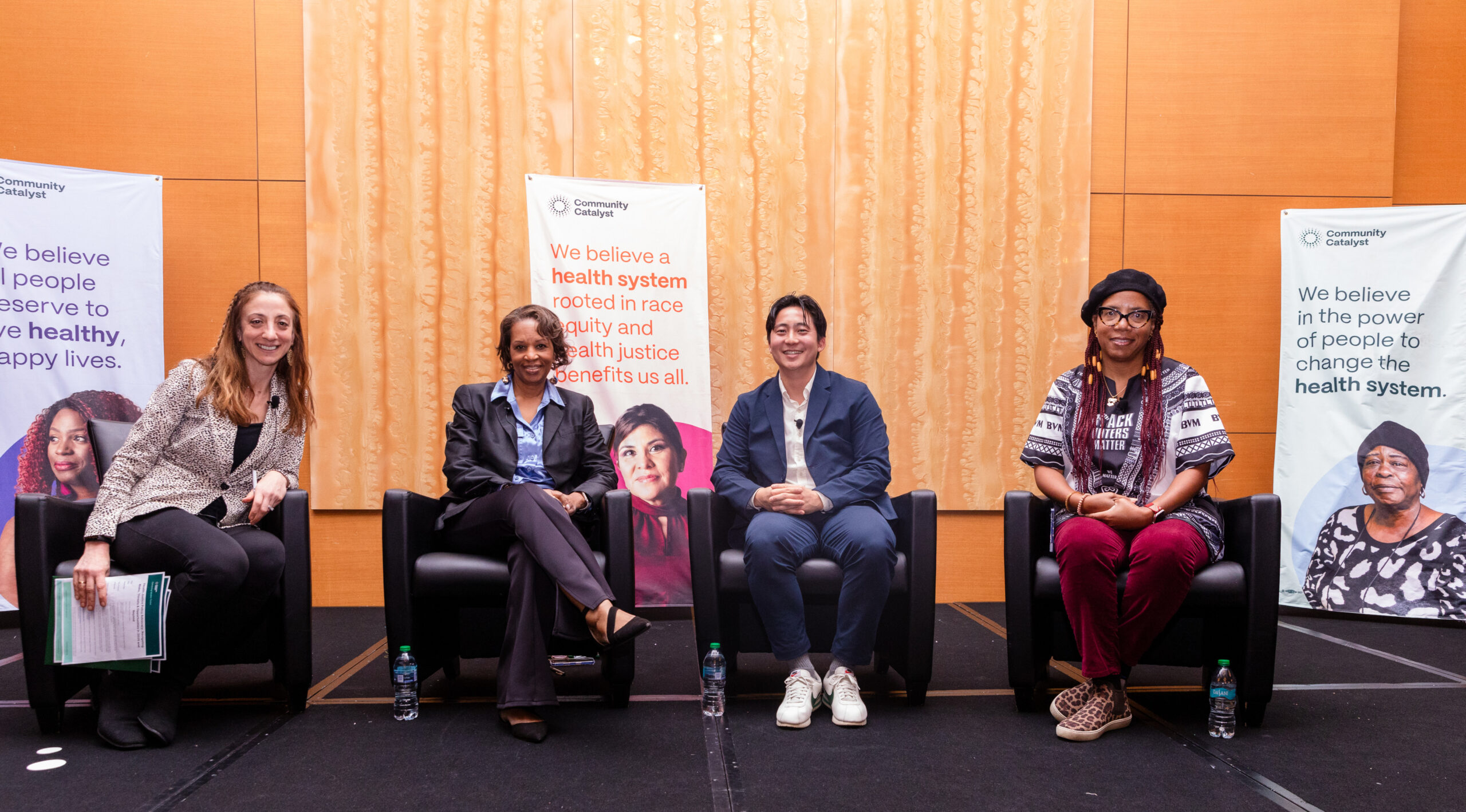 Four smiling people seated in dark chairs in front of a tan backdrop. From left, a woman with long, light brown hair wearing a buttoned gray suit coat and dark pants. Then a woman with short black hair, a black suit with a blue shirt. Then a man with medium length hair, a blue suit, white shirt and white sneakers. Finally a woman with long, braided red hair wearing a flat black hat, a black and white patterned shirt and red pants.