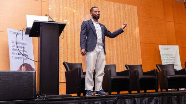 A man in a casual suit addresses an audience on stage