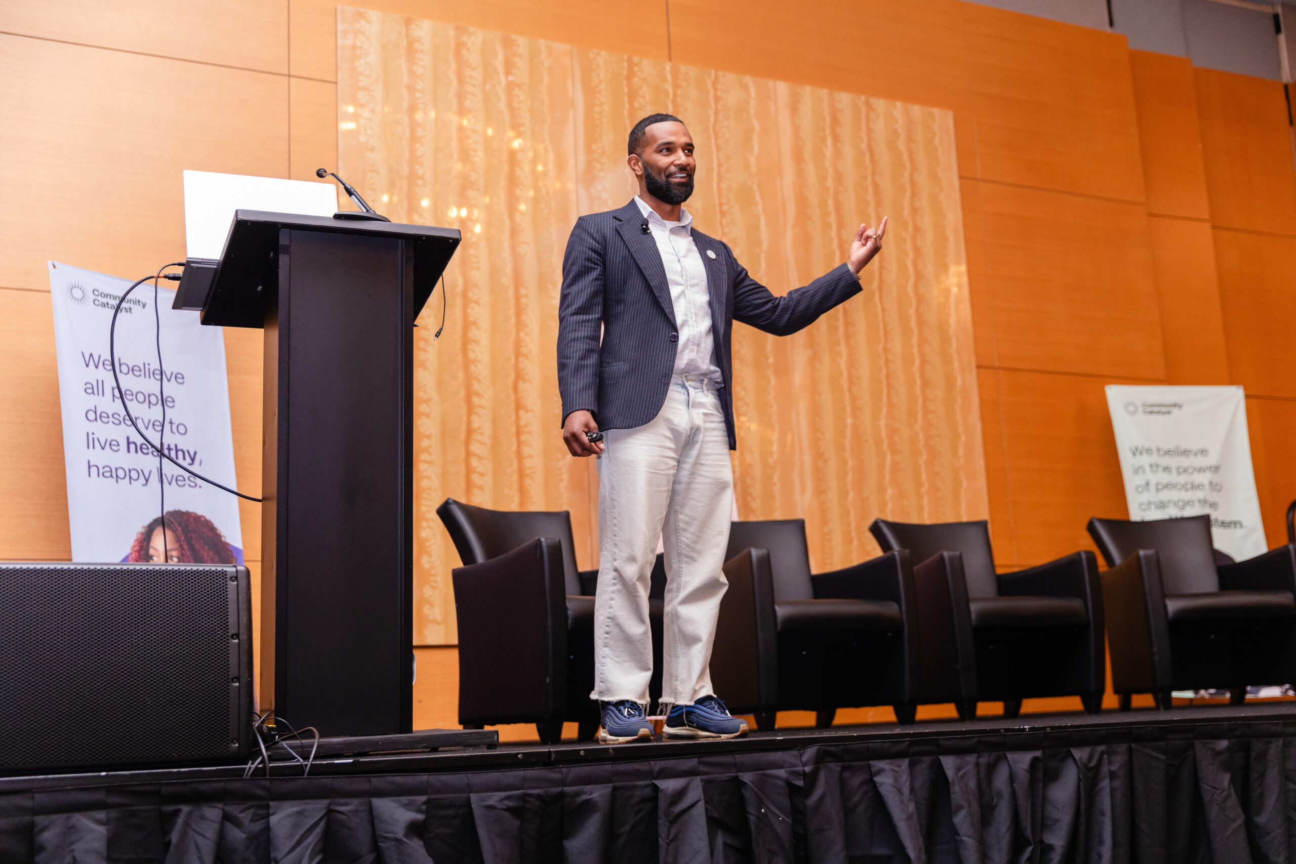 A man with short hair and a beard stands on a stage next to a podium and in front of four brown chairs. He is wearing an open blazer over a white shirt, cream pants and blue tennis shoes.