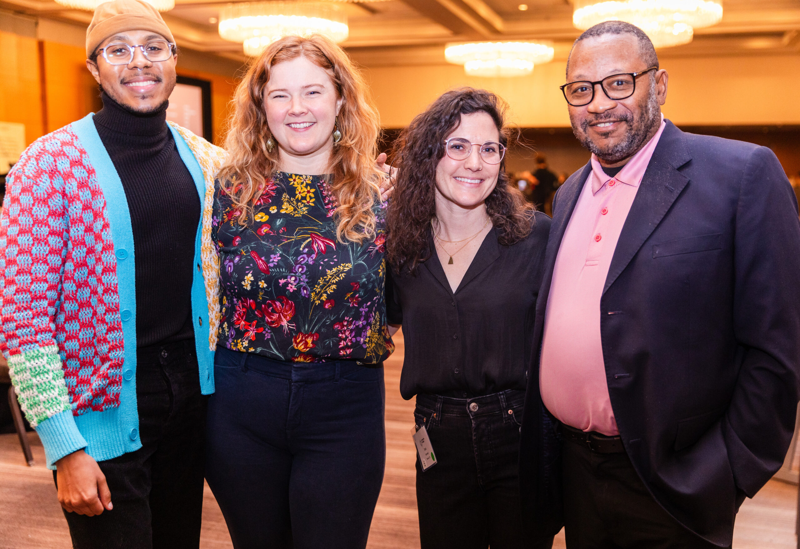 Four people smiling with arms around each other. From left, a man with glasses wearing a tan beanie and a red, blue, white a green open knit sweater over a black shirt and pants. Then a woman with long red hair, a flower-patterned shirt and dark jeans. Then a woman with long black gair and glasses with a black shirt and black jeans. Finally, a man with glasses and a short dark and grey hair and beard wearing a dark suit coat over a pink shirt and dark pants.