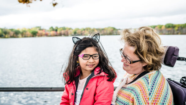 A mother and daughter smile in front of a lake. The mother is seated in a wheelchair with her daughter seated on her lap.