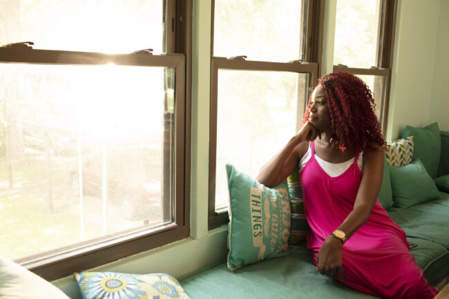 Woman sits on a green couch and looks out a living room window during the daytime. She is wearing a pink and white dress, a gold watch, and has red hair.