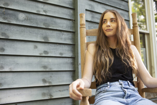 A woman sits in a wooden chair on a front porch. She is wearing a black top and blue jeans.