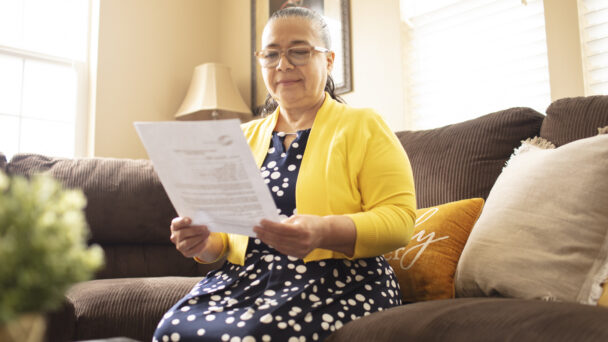 A mother named Angelina sits on the sofa and reads a document. She is wearing a yellow cardigan over a patterned long dress. The room is filled with natural light from a window.