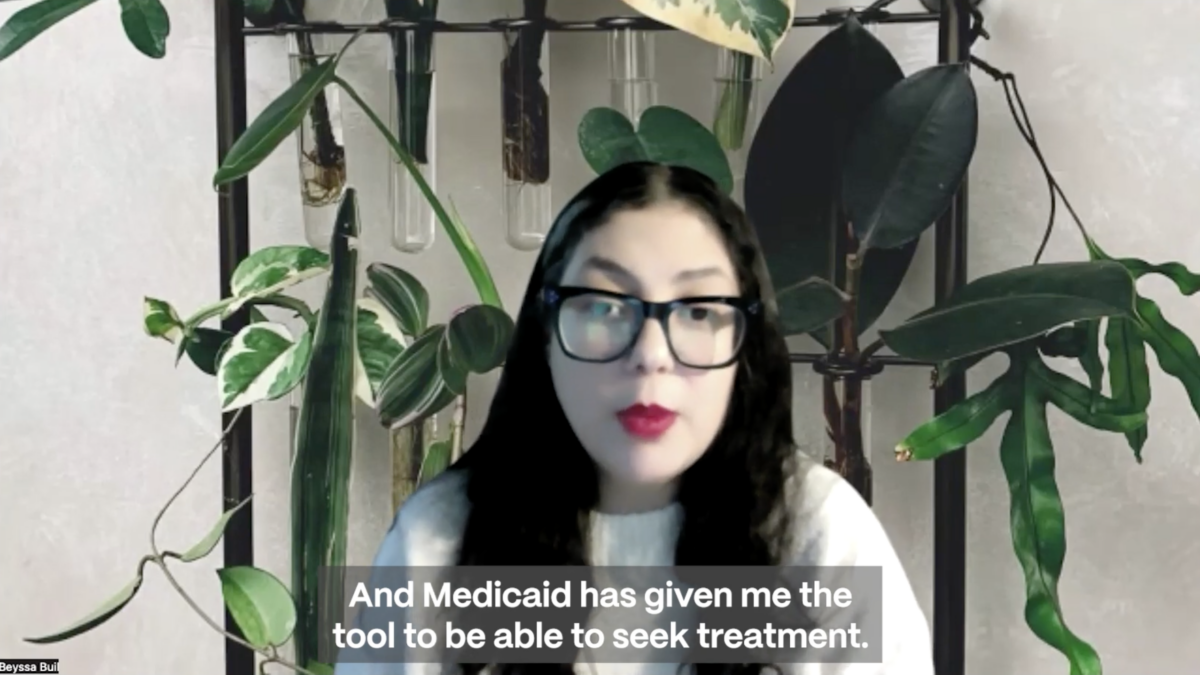 A woman with black hair speaks in front of a background of plants. She is wearing black glasses and red lipstick and a white sweater. A caption at the bottom of the screen reads as "And Medicaid has given me the tool to be able to seek treatment."