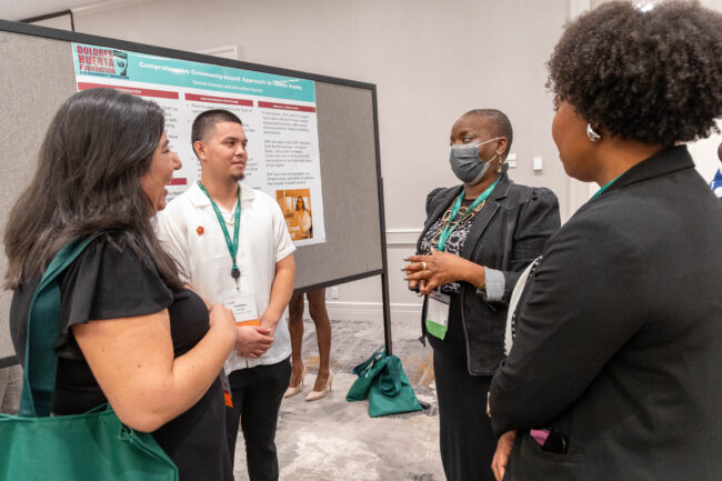 Four community based health advocates speak in a circle. One person is wearing a mask while standing in front of a poster about community-based approaches to health equity