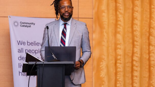 Brandon G. Wilson speaks at the 2024 Community Catalyst Convening. He is standing at a podium in front of a mic and a laptop. Behind him is a sign that reads, "We believe all people deserve to live a happy, healthy life."