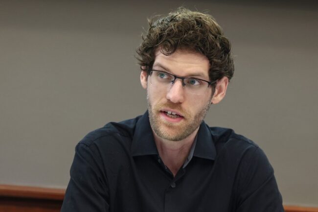 Jason speaks in a conference room. He is wearing glasses and a black shirt.