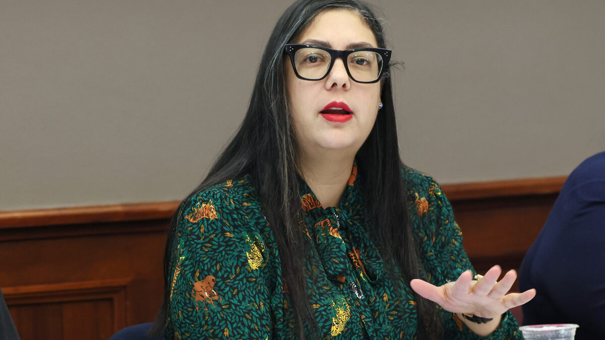 A woman with long black hair, black glasses, and red lipstick speaks at an event. She is wearing a green blouse.