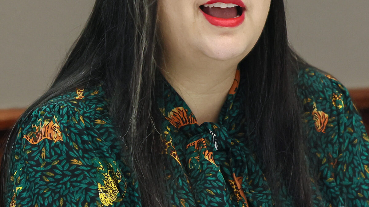 A woman with long black hair, black glasses, and red lipstick speaks at an event in D.C. She is wearing a green blouse