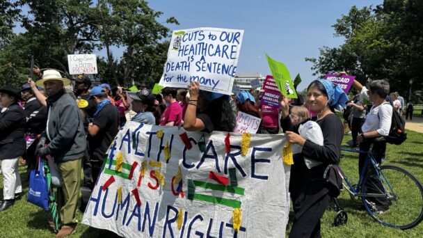 A group of people hold signs at a protest demand no cuts to our care and defend Medicaid and the ACA. The sign reads "cuts to our health care is a death sentence for the many" and "health care is a human right."