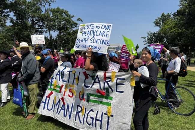 A group of people hold signs at a protest demand no cuts to our care and defend Medicaid and the ACA. The sign reads "cuts to our health care is a death sentence for the many" and "health care is a human right."