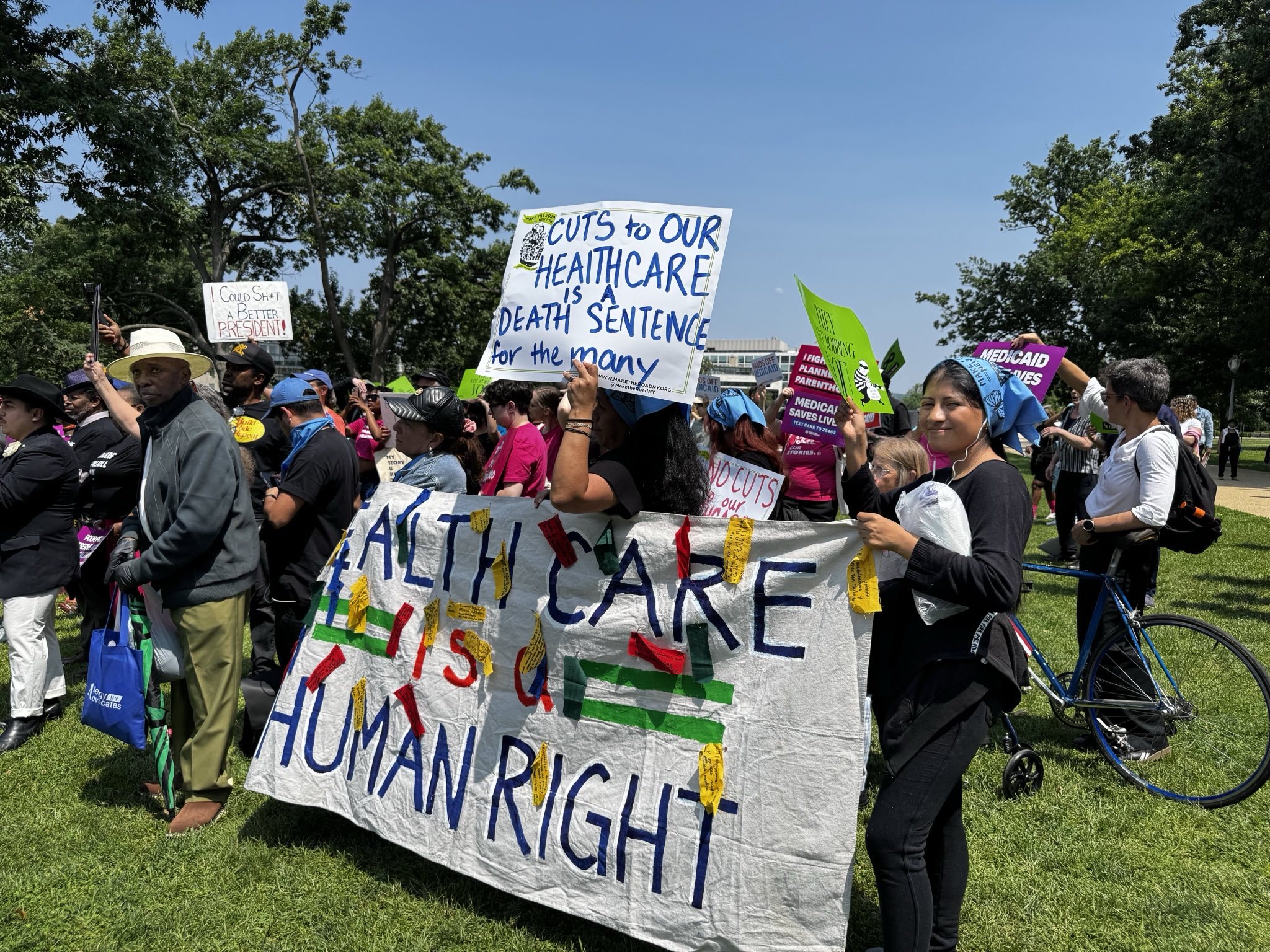A group of people hold signs at a protest demand no cuts to our care and defend Medicaid and the ACA. The sign reads "cuts to our health care is a death sentence for the many" and "health care is a human right."