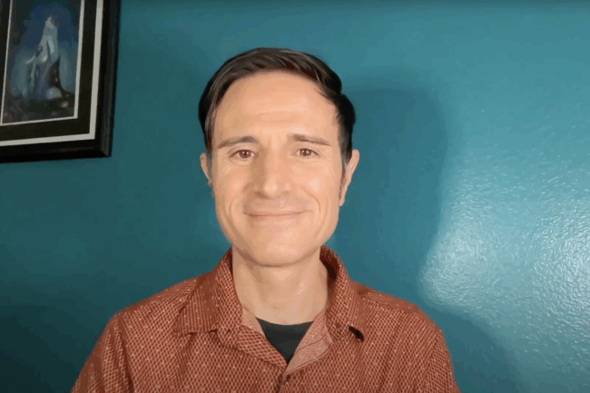 Ricky speaks in front of a blue wall and a framed painting. He has brown hair and is wearing an orange shirt.