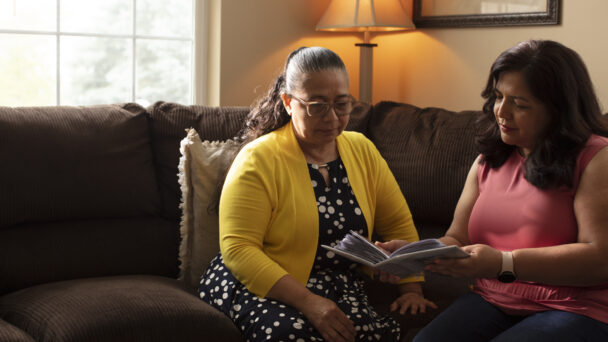 A mother named Angelina and her daughter, Suzy, sit on the sofa and look at a photo album. Angelina wears a yellow cardigan over a patterned long dress. Suzy wears a salmon pink sleeveless blouse and jeans. The room is filled with natural light from a window.